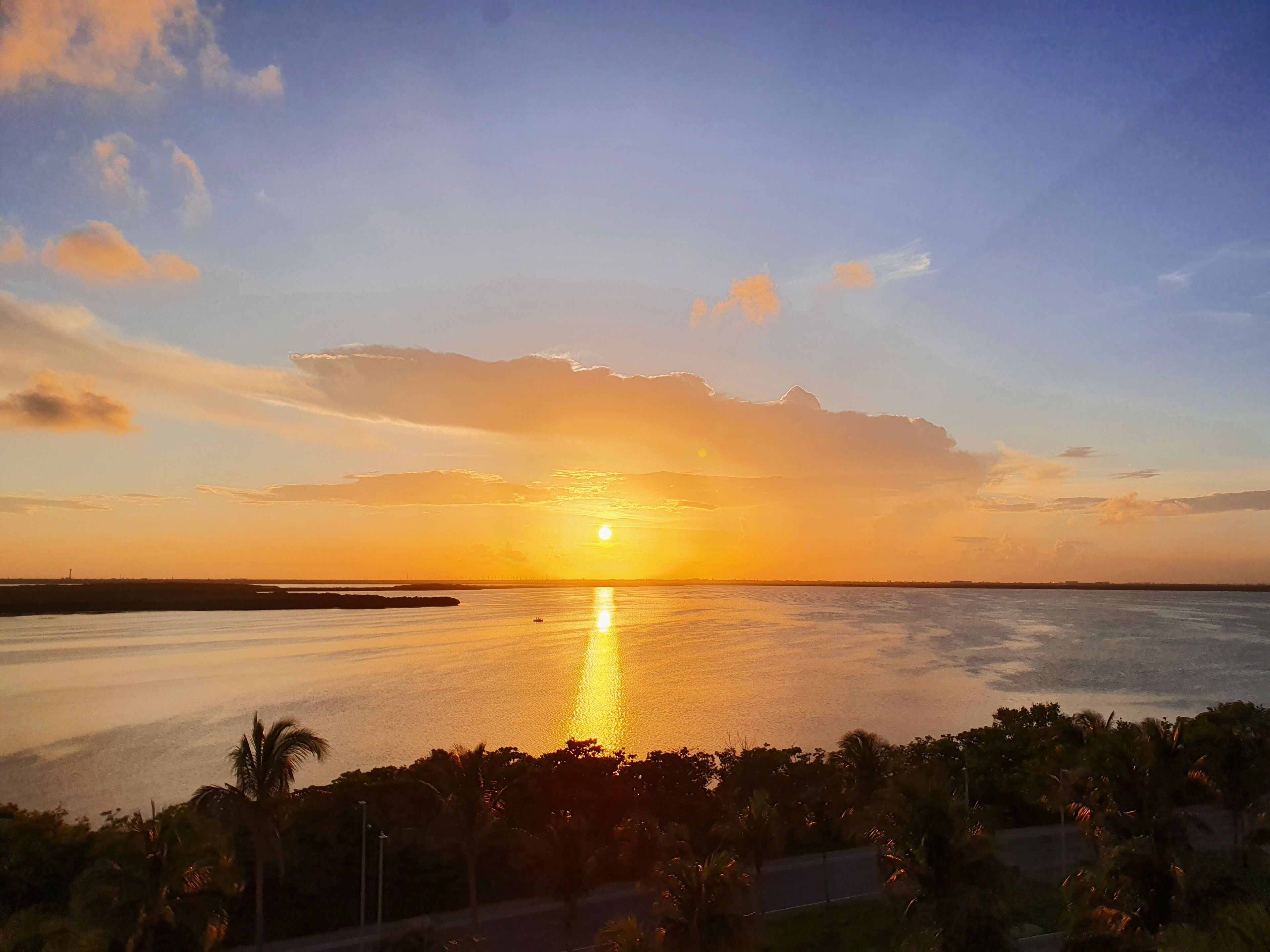 Un atardecer sobre una superficie acuática tranquila, con una orilla lejana y palmeras en primer plano.