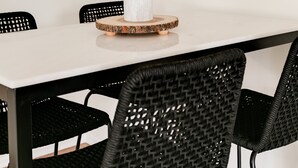 Un'area pranzo con un tavolo bianco, sedie nere intrecciate, uno specchio rotondo con cornice intrecciata e una piccola pianta verde su un vassoio di legno.
