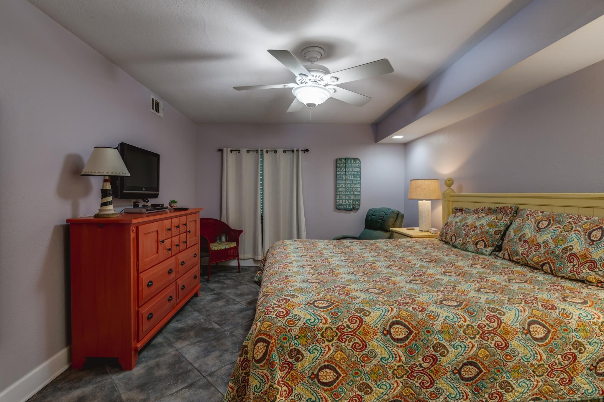 A bedroom with a bed, a television on a dresser, a ceiling fan, and a nightstand with a lamp.