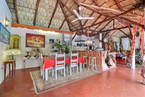 Un coin repas avec une nappe rouge, des chaises blanches, un ventilateur de plafond et un plafond en bois rustique.