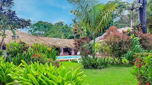 A lush garden with a thatched-roof structure, a swimming pool, and various tropical plants.