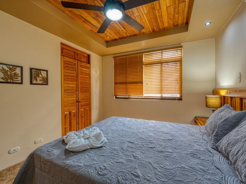 Une chambre à coucher avec un lit, un ventilateur de plafond en bois, des stores aux fenêtres et une lampe fixée au mur.