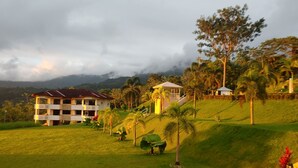 A resort with multiple buildings, including a large house with a red roof and a smaller structure with a white roof. There are palm trees and a grassy hill in the foreground. - Private Garden, Walk Out to Community Pool, Beautiful 1 Bedroom 1 Bath Condo (San Buenas)