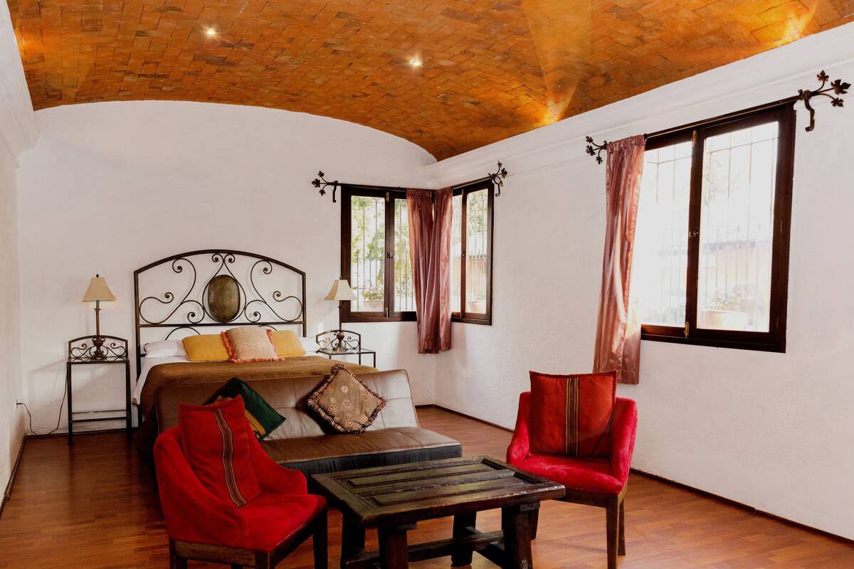 A bedroom with a wooden ceiling, a bed with a headboard, a wooden table, two chairs, and a window with curtains.