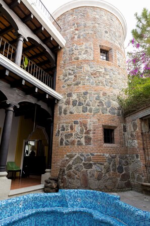 Ein Steinturm mit Balkon, ein Schwimmbecken mit blauen Mosaikfliesen und ein Stuhl davor.