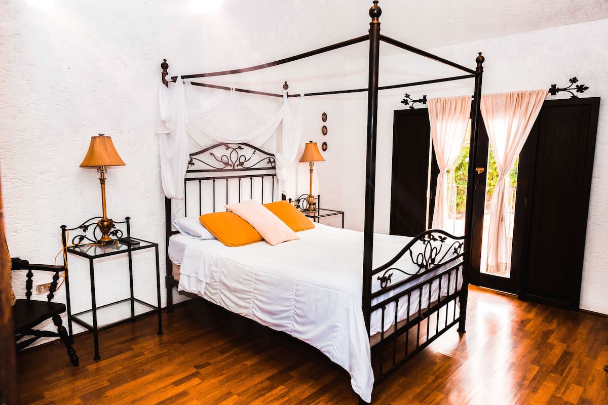 A four-poster bed with white bedding and orange pillows, two bedside tables with lamps, a wooden floor, and a window with curtains.