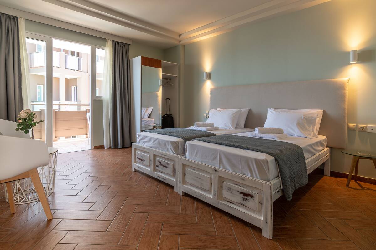 A hotel room with a double bed, a small table, a chair, a mirror, and a view of the outside balcony.