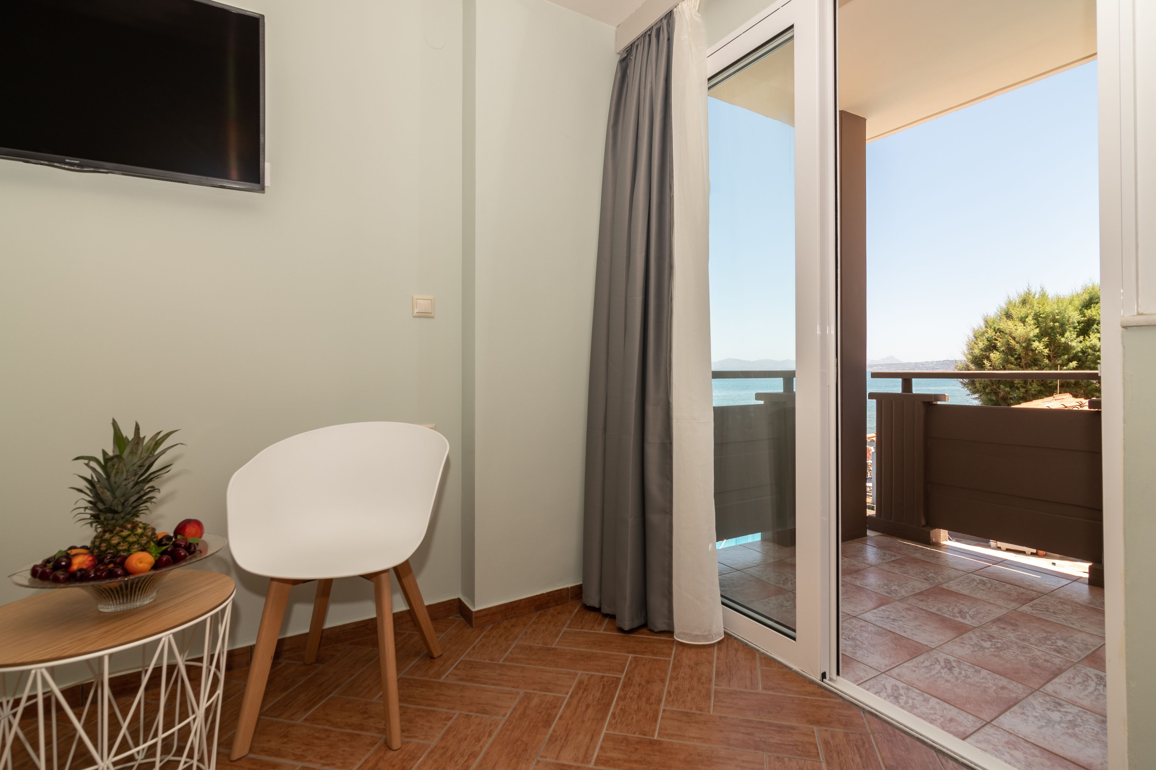 A balcony with a view of the sea, a white chair, a small table with fruit, and a TV mounted on the wall.