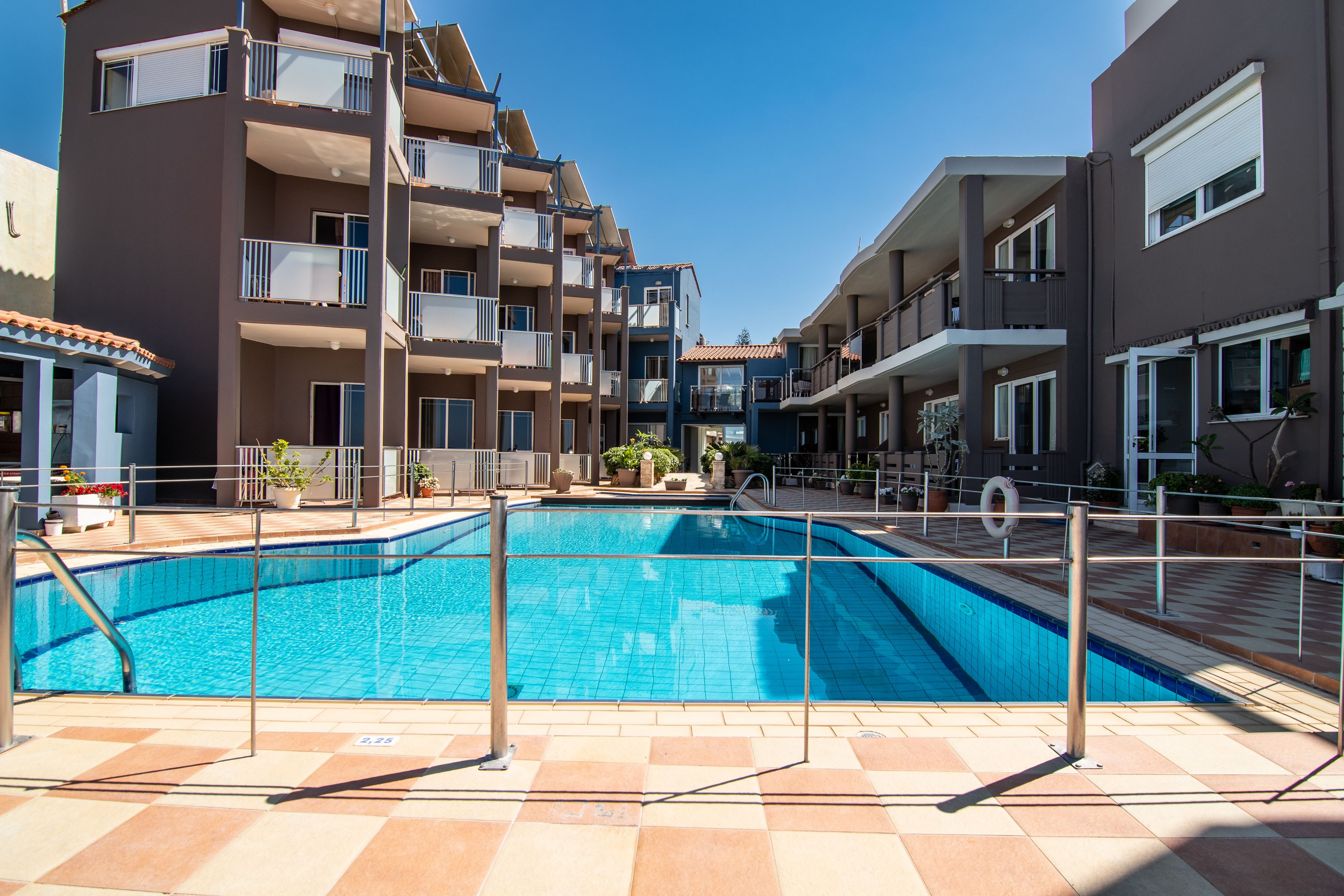 A swimming pool surrounded by multi-story buildings with balconies, a tiled deck, and potted plants.