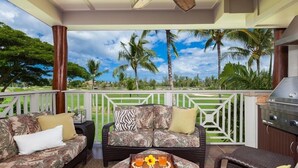 A covered outdoor seating area with a sofa and chairs, a grill, and a view of palm trees and greenery. - Hilton Pool Pass for stays 2026. Golf Discounts. Waikoloa Beach Villas L22 (Waikoloa Beach Resort)