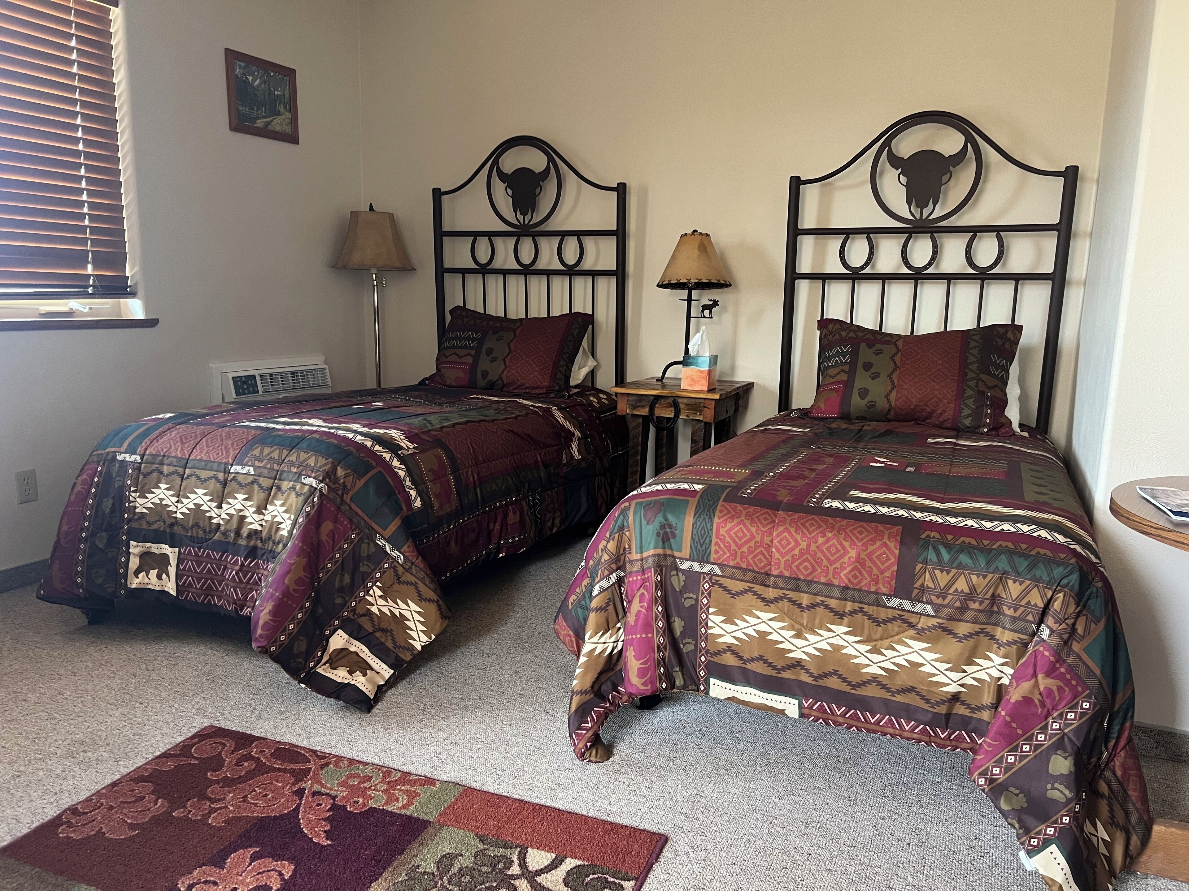 Two twin beds with patterned quilts, a small bedside table with a lamp, and a decorative rug on the floor.