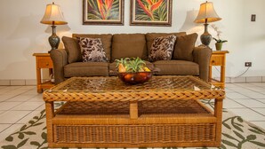 Uma sala de estar com um sofá marrom, duas mesas laterais com abajures, uma mesa de centro de vidro com uma tigela de frutas e dois quadros emoldurados na parede.