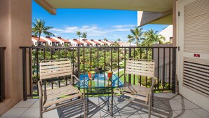 Uma varanda com mesa e cadeiras de vidro, vista para um resort com palmeiras e piso de azulejos.