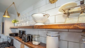 A kitchen with white tiled walls, wooden shelves, and various kitchen items including a colander, jars, and a kettle.
