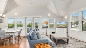 A spacious living area with a white ceiling, large windows with blinds, a grey sofa, a dining table with chairs, and a wooden side table.