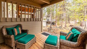 A covered outdoor seating area with wicker furniture and green cushions, overlooking a forested area. - ASHW05 - Charming Home, A/C, Hot Tub,  Bikes (Sunriver)