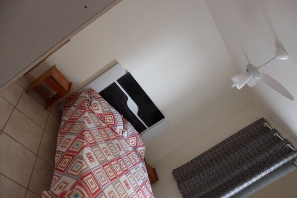A room with a bed, a ceiling fan, a black and white poster on the wall, and a patterned curtain.