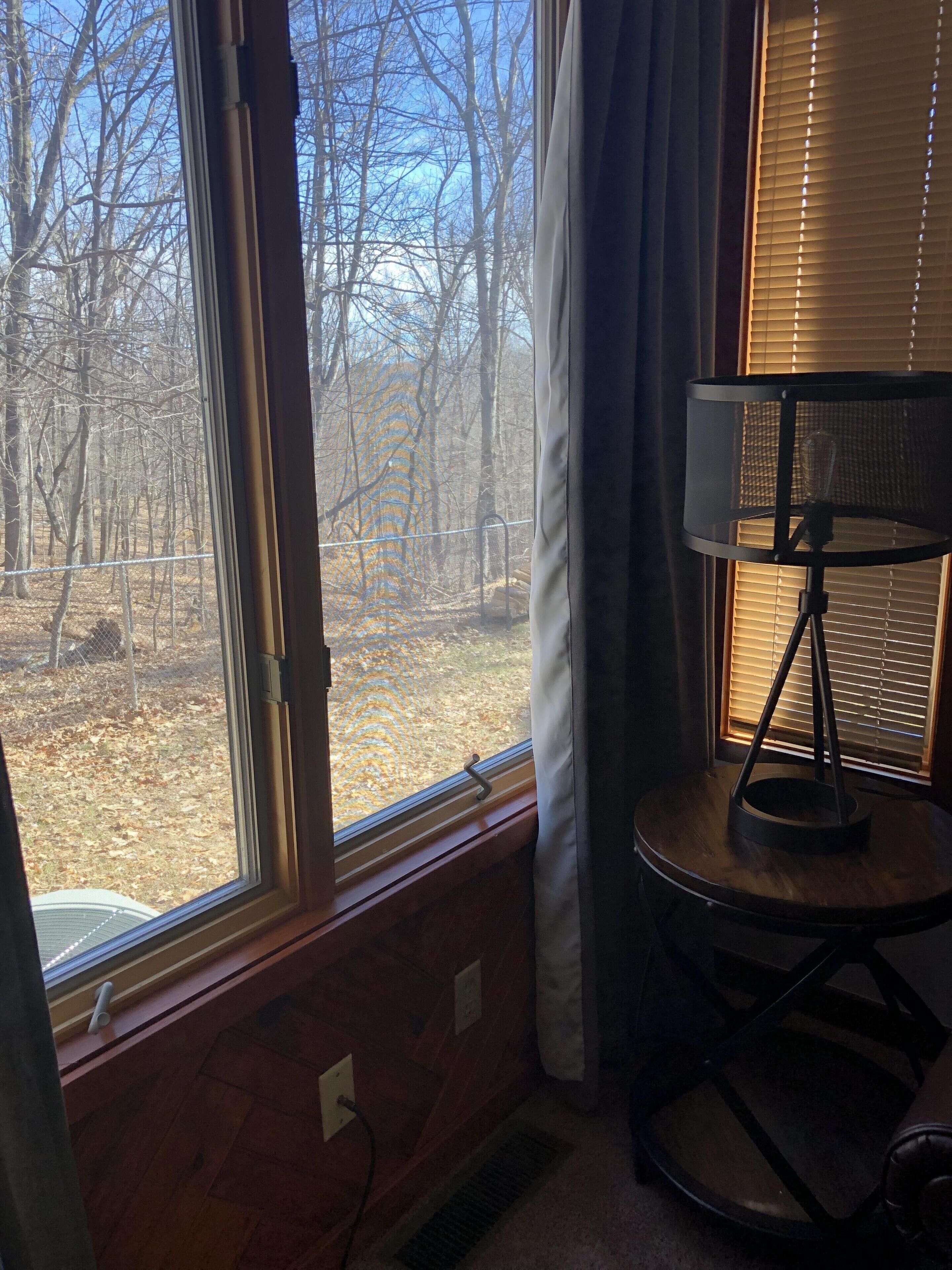 A room with a large window offering a view of a wooded area. There is a small round table with a lamp on it.