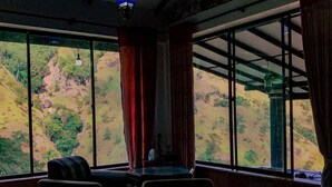 Quarto com uma grande janela com vista para uma paisagem montanhosa. Há duas cadeiras e uma pequena mesa.