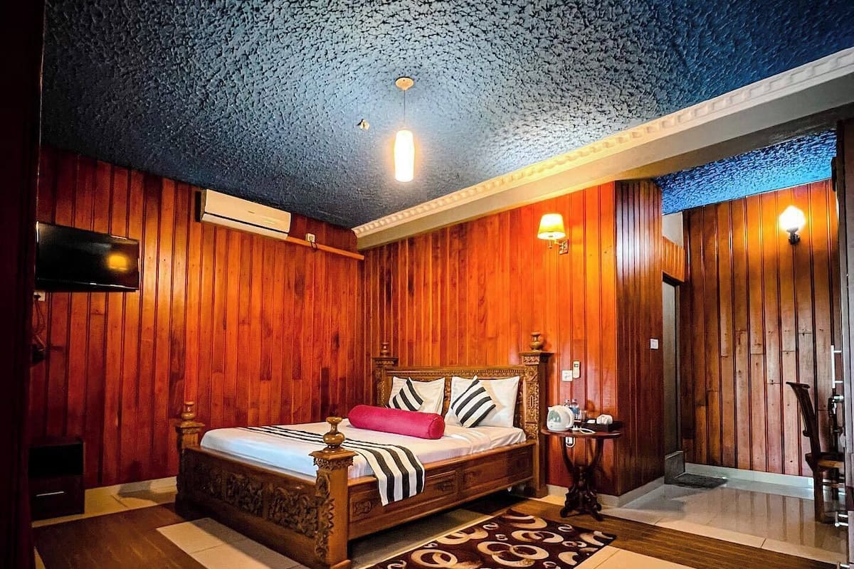 Quarto com painéis de madeira, uma cama, uma mesa pequena e uma televisão fixada na parede.