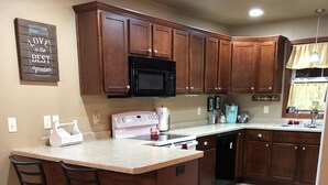 Cocina con armarios de madera, microondas, estufa y fregadero. Hay dos taburetes de bar y una encimera.