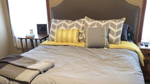 Una cama bien tendida con una colcha gris y amarilla, una almohada estampada en gris y blanco, y un cabecero gris.