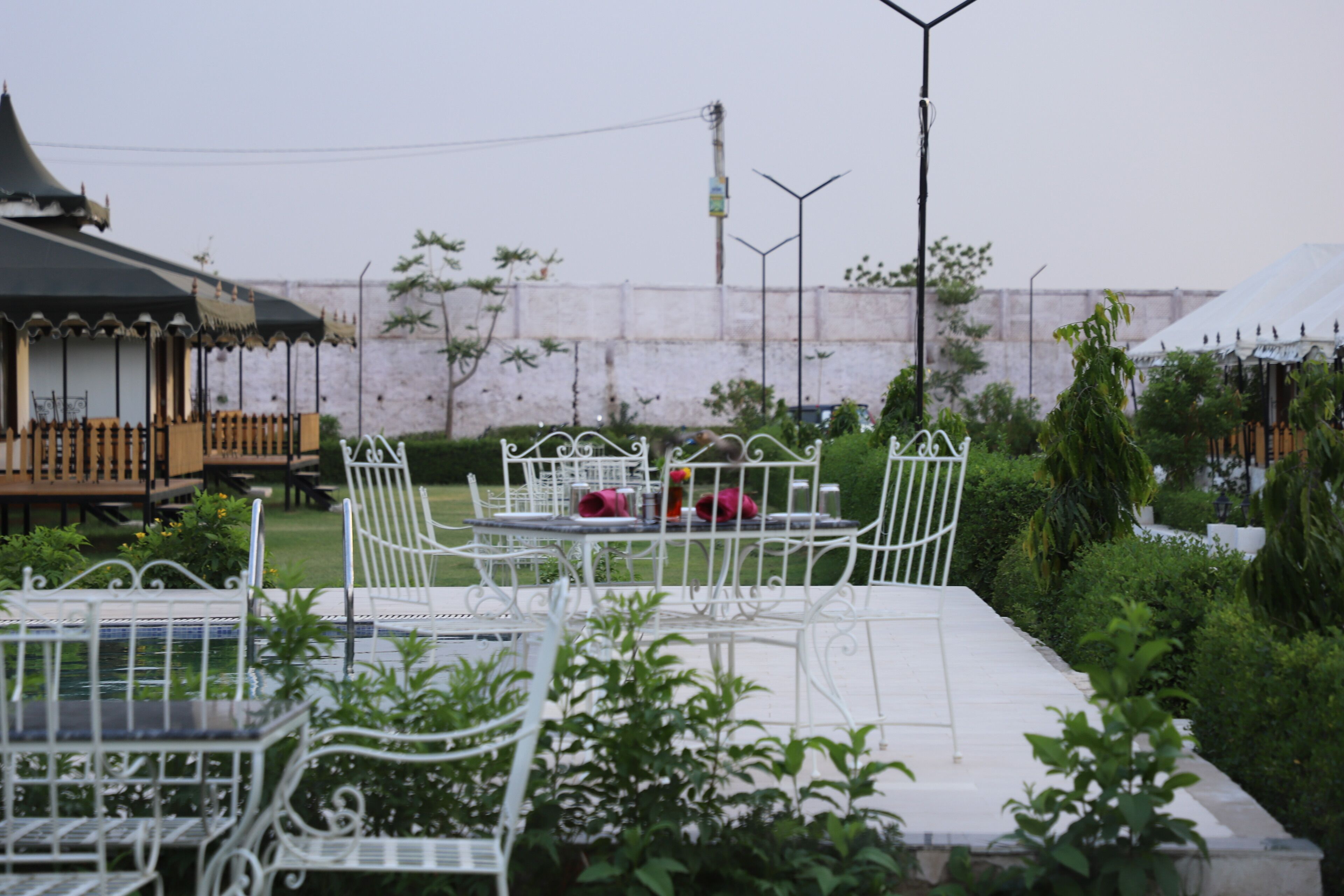 Outdoor seating with metal chairs and tables, surrounded by greenery and a white wall in the background.