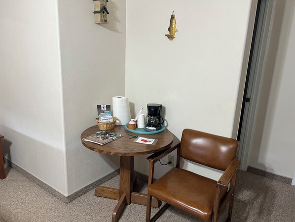 Una pequeña mesa de madera con una cafetera, una cesta y una revista. Un sillón de cuero marrón está al lado de la mesa.