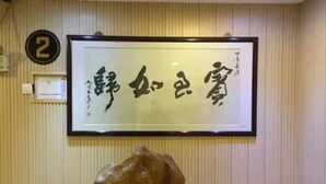 Una obra de caligrafía enmarcada en una pared, un letrero con el número 2 y una escultura de roca sobre una mesa.