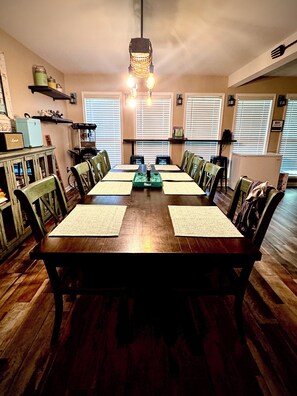 Une salle à manger avec une table en bois, des chaises, un lustre et une vue dégagée sur l’extérieur grâce à de grandes fenêtres.