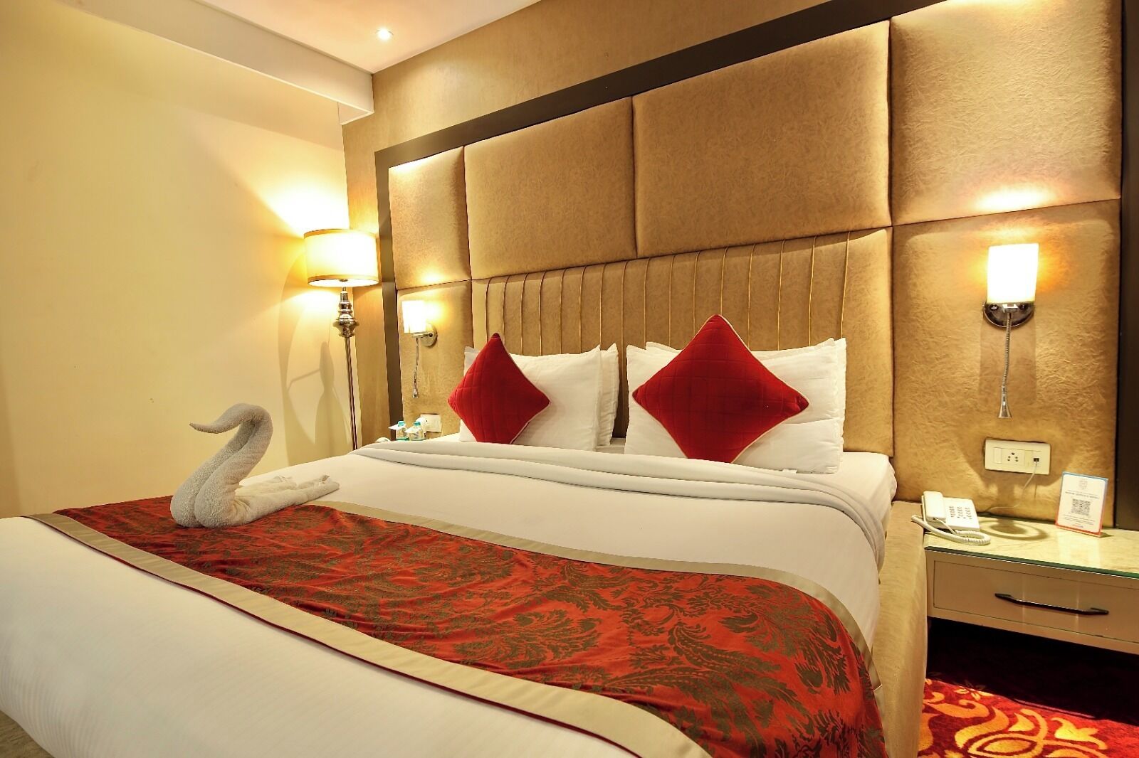 Une chambre d’hôtel avec un grand lit, une couvre-lit rouge et or à motifs, deux oreillers rouges et une serviette en forme de cygne, ainsi qu’un téléphone sur la table de chevet.