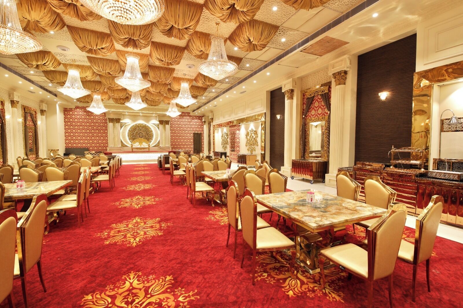 Une salle de banquet dotée de nombreuses tables rondes et de chaises, d’un tapis rouge et de lustres somptueux.