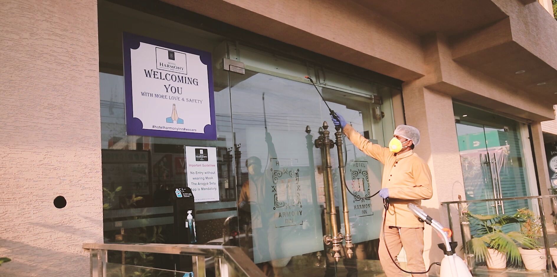 A person spraying a disinfectant outside a building with a sign reading "WELCOME YOU WITH MORE LOVE & SAFETY."