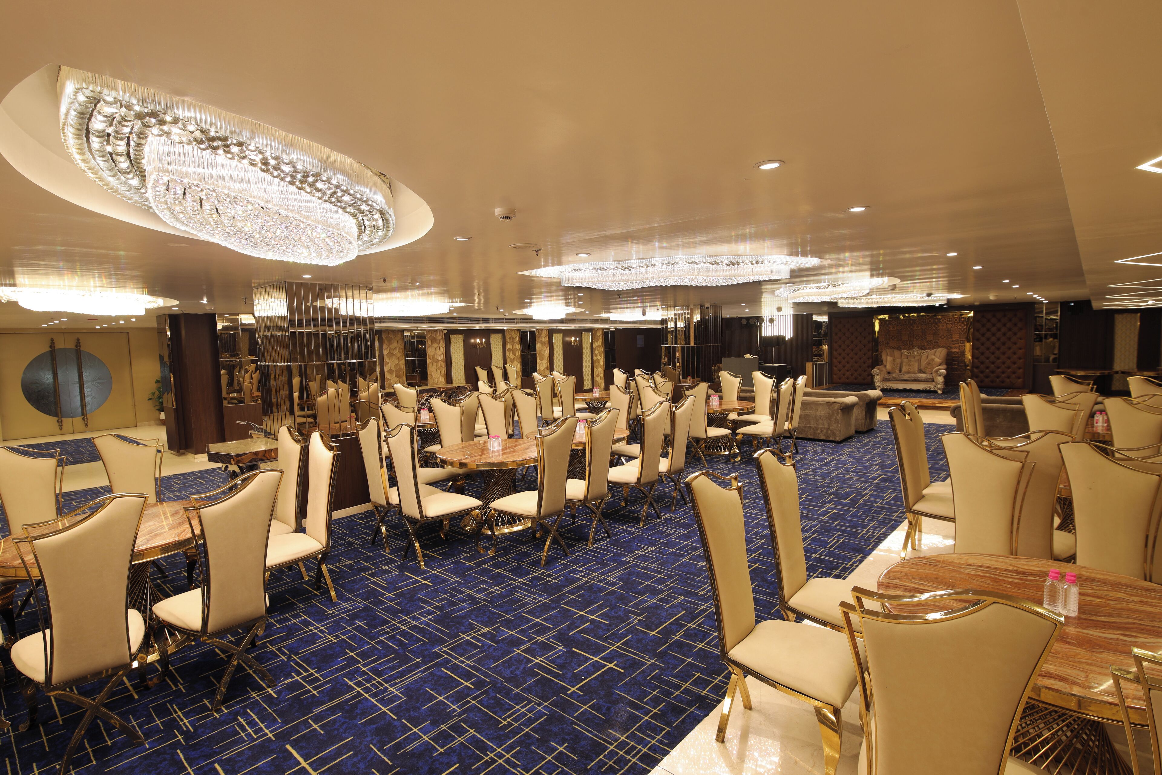 Amplio salón comedor de hotel con varias mesas y sillas, alfombra de diseño azul y una gran lámpara de araña.