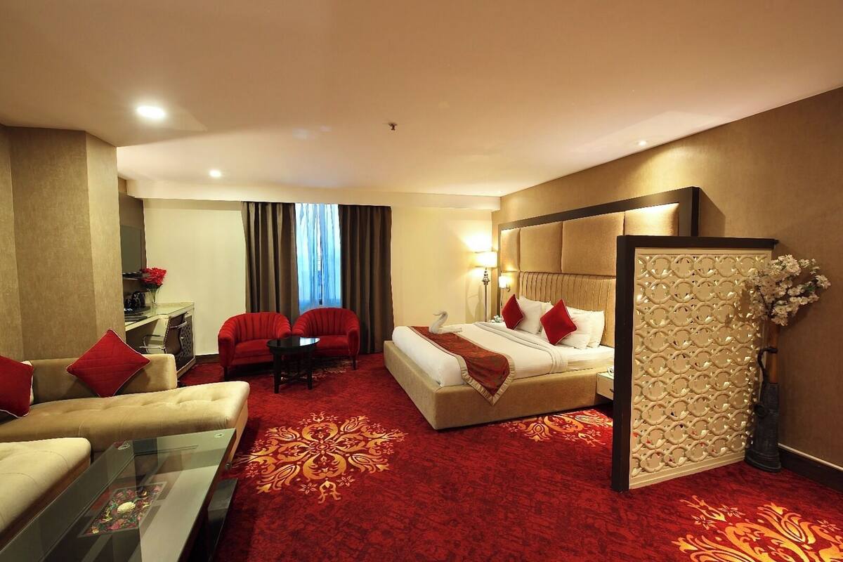 A hotel room with a bed, two armchairs, a sofa, a glass coffee table, and a decorative wall panel.