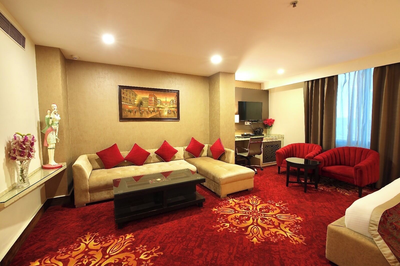 A living room with a beige sofa, red cushions, a red armchair, a coffee table, a TV, and a red carpet.