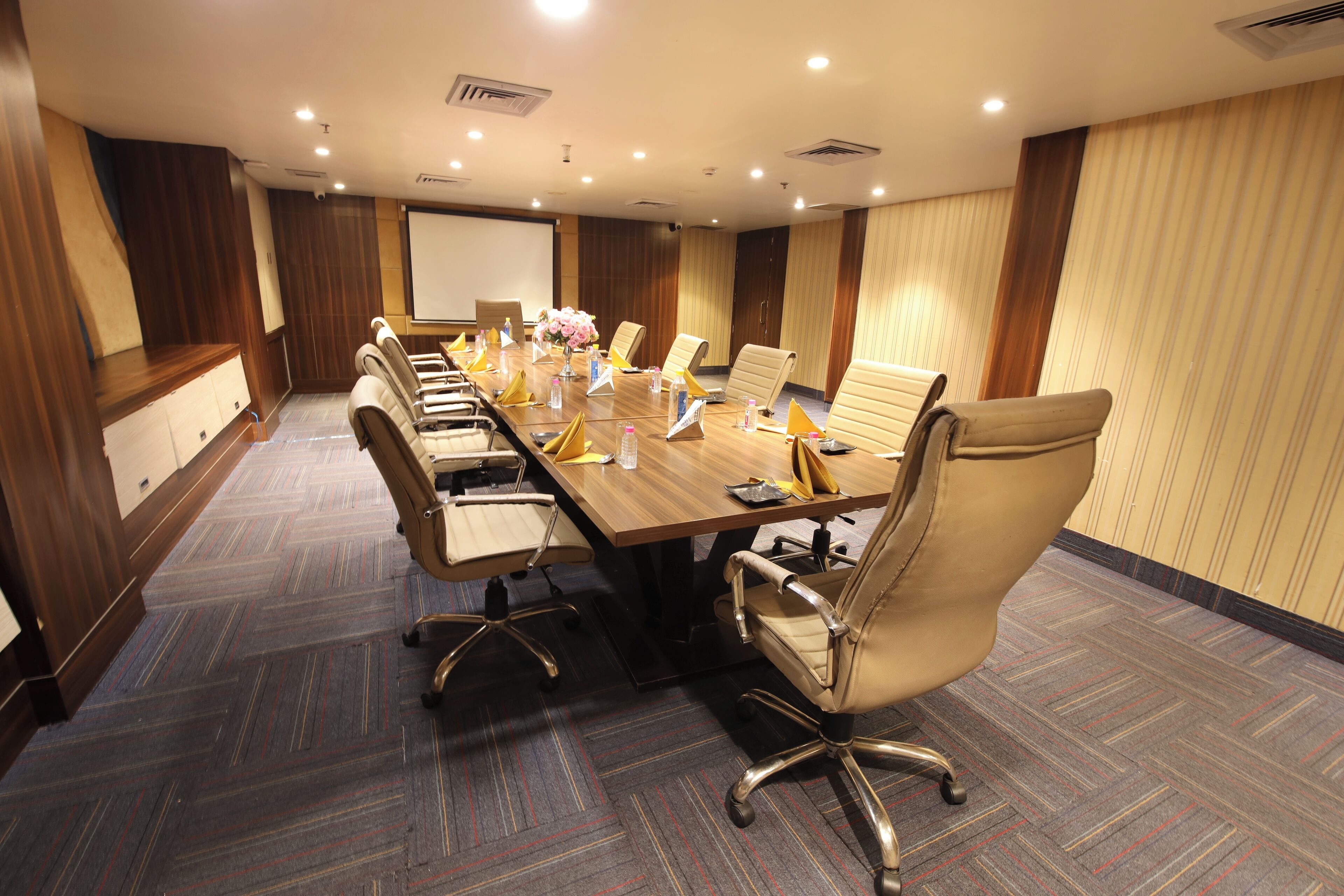 Un salón de conferencias con una larga mesa, varias sillas, una pantalla de proyector y un arreglo floral.