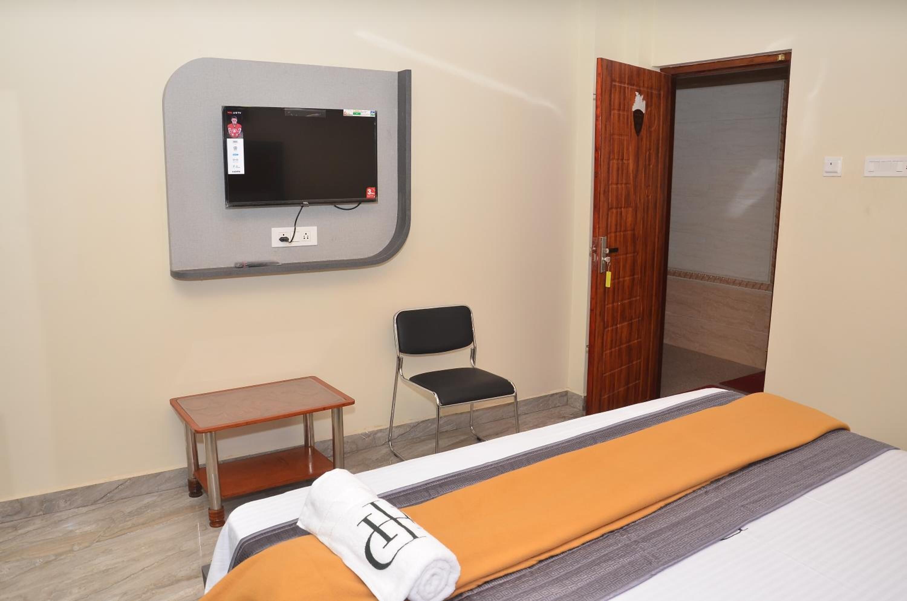 A hotel room with a bed, a small table, a chair, a TV mounted on the wall, and a door.