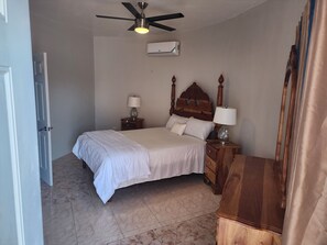A bedroom with a bed, a nightstand with a lamp, a ceiling fan, and an air conditioning unit.
