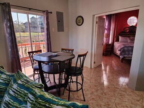 Un área de comedor con una mesa y sillas, vista a un balcón a través de una puerta corrediza, y una cama en la habitación contigua.