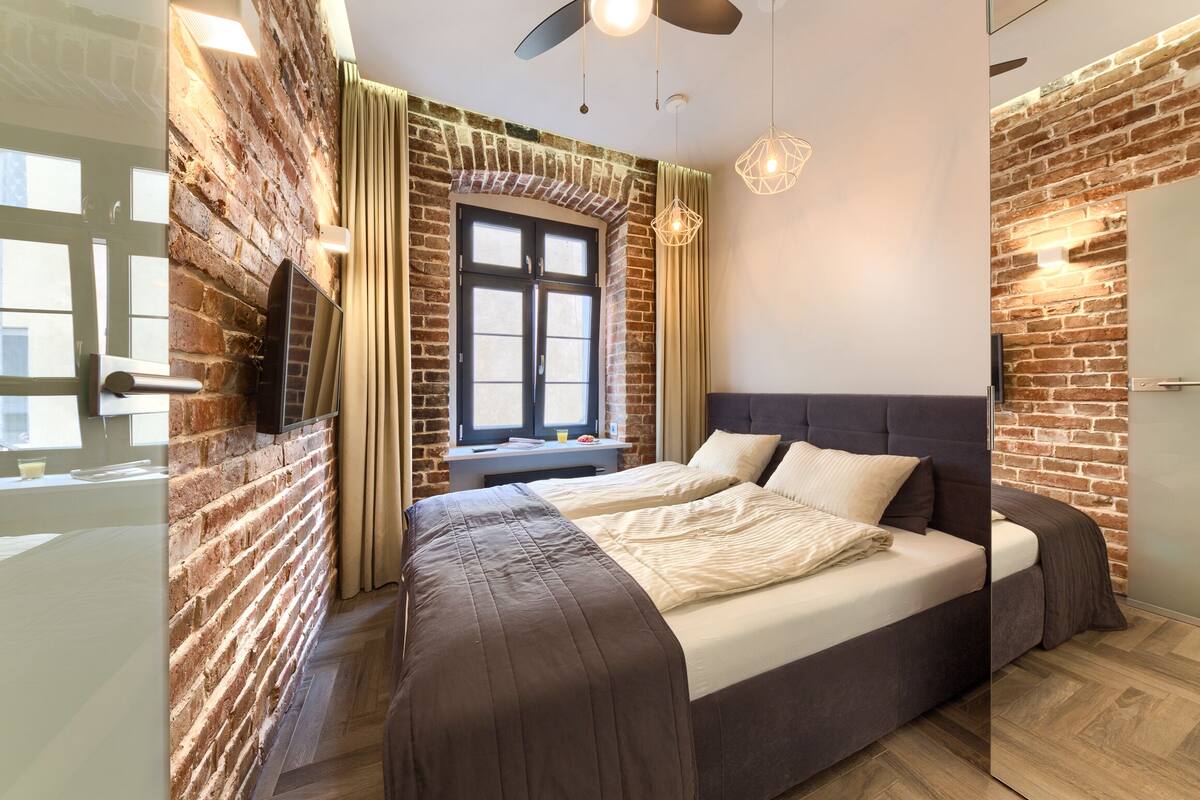 A modern bedroom with a brick wall, a large bed, a ceiling fan, and a window with curtains.