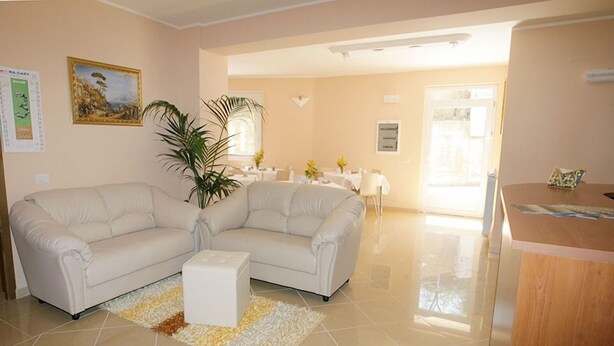 A living room with a beige sofa, a coffee table, and a potted plant. There is a dining area in the background with a table and chairs.