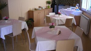 A dining area with tables set for four, a kitchen in the background, and a window providing natural light.
