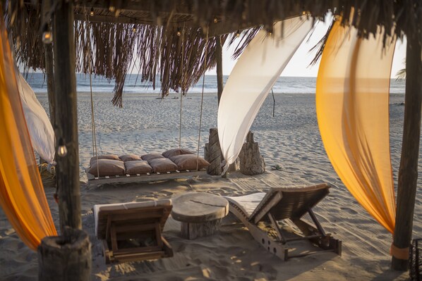 Un salón junto a la playa con techo de paja, asientos acolchados y mobiliario de madera.