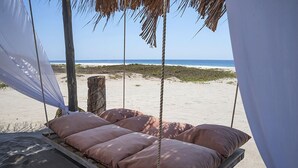 Un hamac de plage agrémenté de coussins, une plateforme en bois et une balançoire en corde, le tout installé sur une plage avec des palmiers en arrière-plan et une vue sur l’océan.
