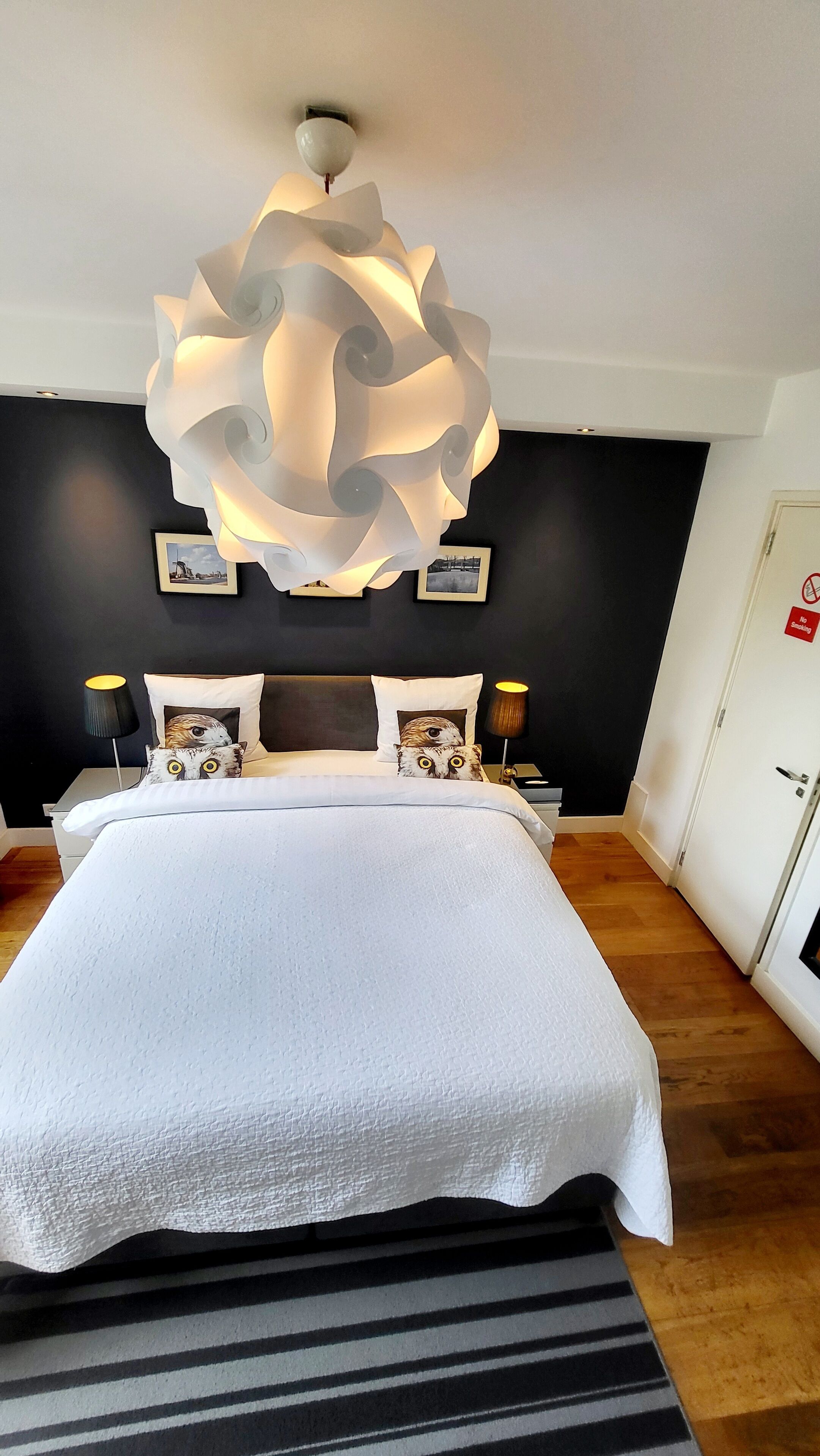 A modern bedroom with a large bed, a unique ceiling light fixture, and a 'No Smoking' sign on the door.