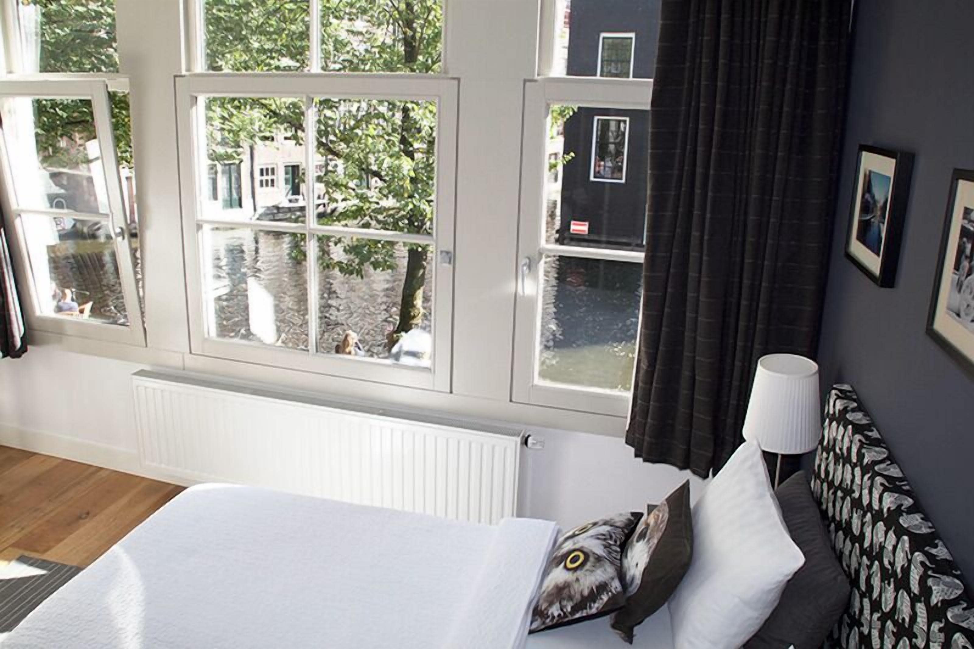 A hotel room with large windows, a bed, a bedside lamp, and a view of a canal.