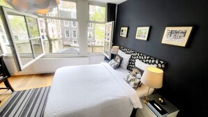Une chambre avec un grand lit, une fenêtre donnant sur des immeubles, une table de chevet avec une lampe et trois tableaux encadrés accrochés au mur.