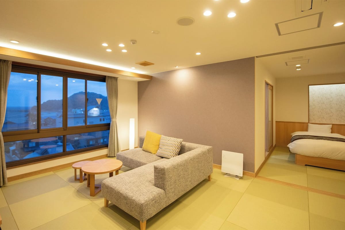 A hotel room with a sofa, a small table, a bed, and a large window offering a view of the sea.