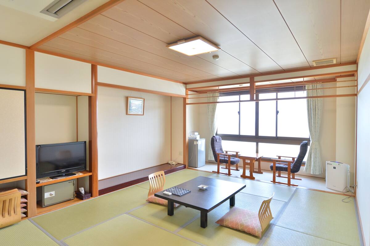 A traditional Japanese room with tatami flooring, a television, a desk, and a view of the outside through large windows.